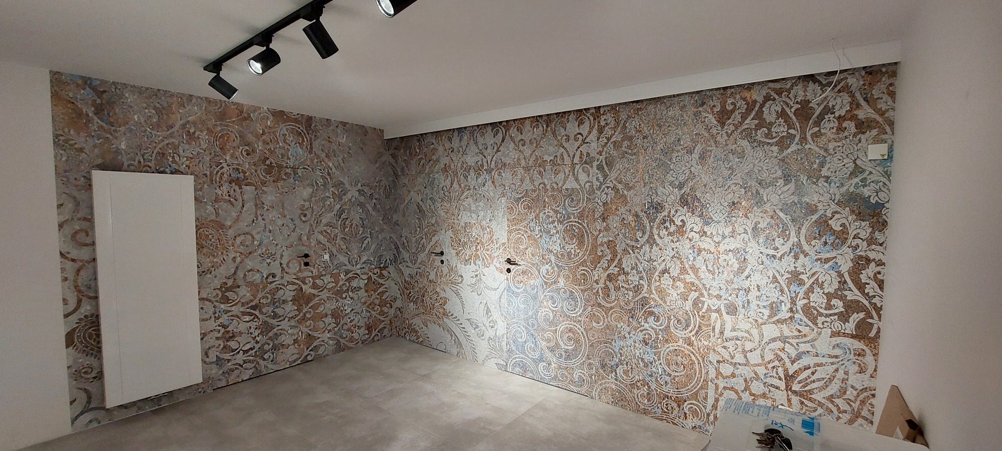 Wykończona łazienka z dekoracyjnymi, wzorzystymi płytkami ściennymi w odcieniach brązu i szarości, jasne płytki podłogowe, biały grzejnik panelowy i czarne reflektory sufitowe.