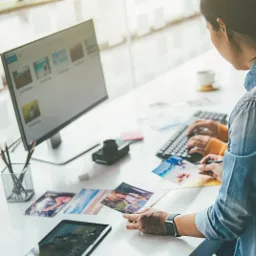 Osoba przy biurku edytuje zdjęcia na komputerze i tablecie, przeglądając wydrukowane fotografie z motywami plażowymi.