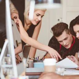 Zespół projektantów analizuje dokumentację na biurku z komputerem, laptopem, kawą i przyborami biurowymi w jasnym, nowoczesnym biurze.