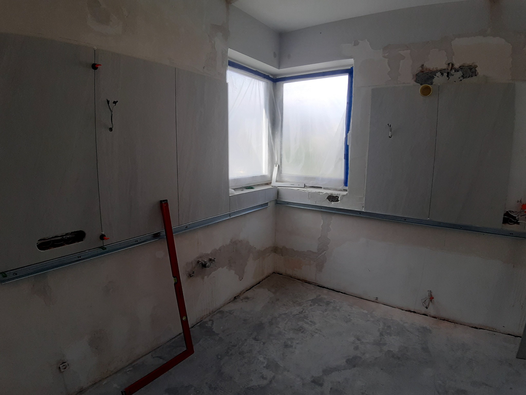 Wnętrze w trakcie remontu: ściany częściowo pokryte płytami, okno zabezpieczone folią, widoczny poziomica oparta o ścianę, metalowe profile zamontowane na ścianach, przygotowane otwory na instalacje.
