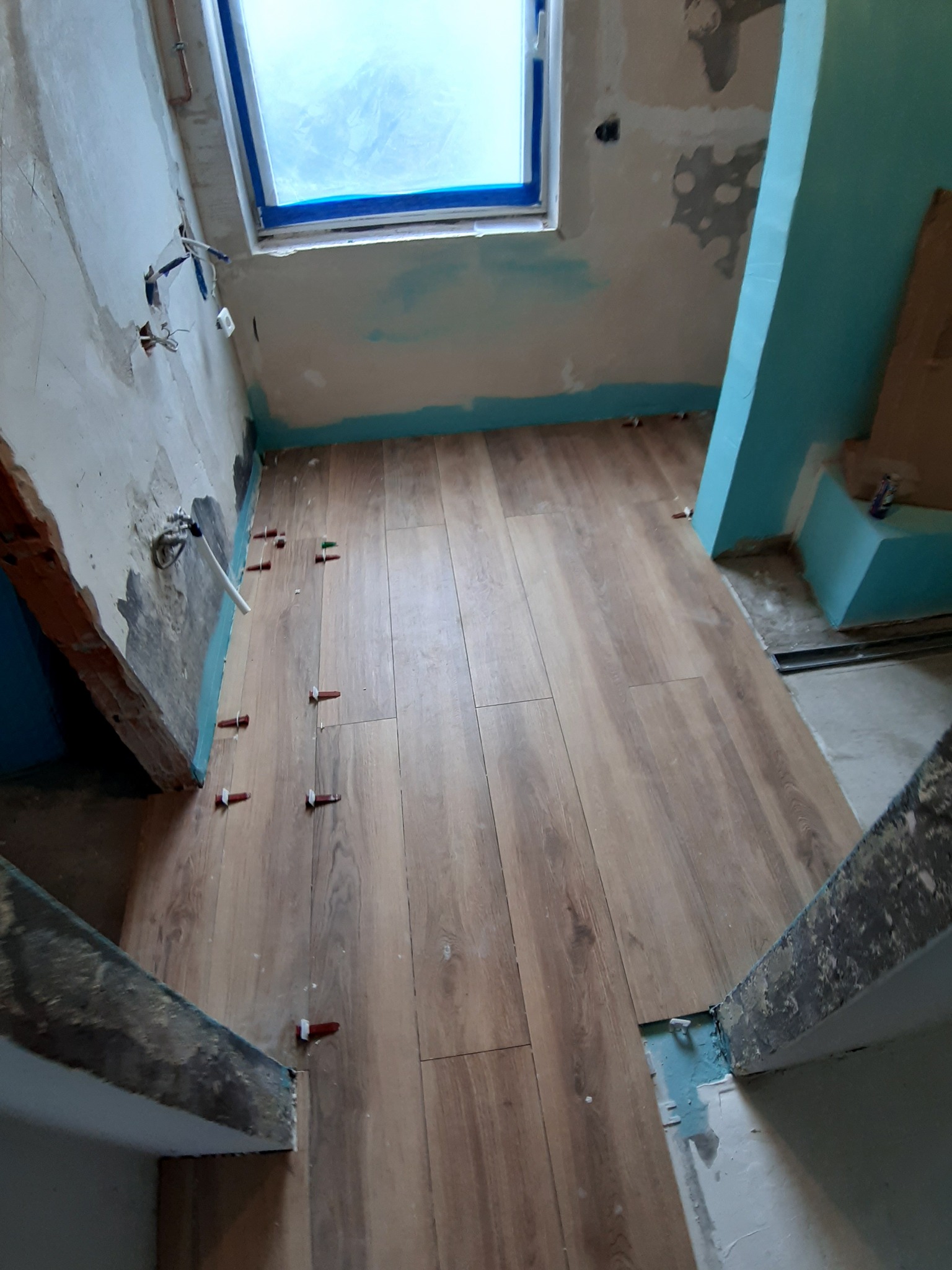 Układanie paneli podłogowych w trakcie remontu łazienki, widoczne dystanse poziomujące, surowe ściany i instalacja elektryczna.