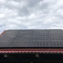 Sześć paneli słonecznych na czerwonym, falistym dachu budynku gospodarczego pod pochmurnym niebem.