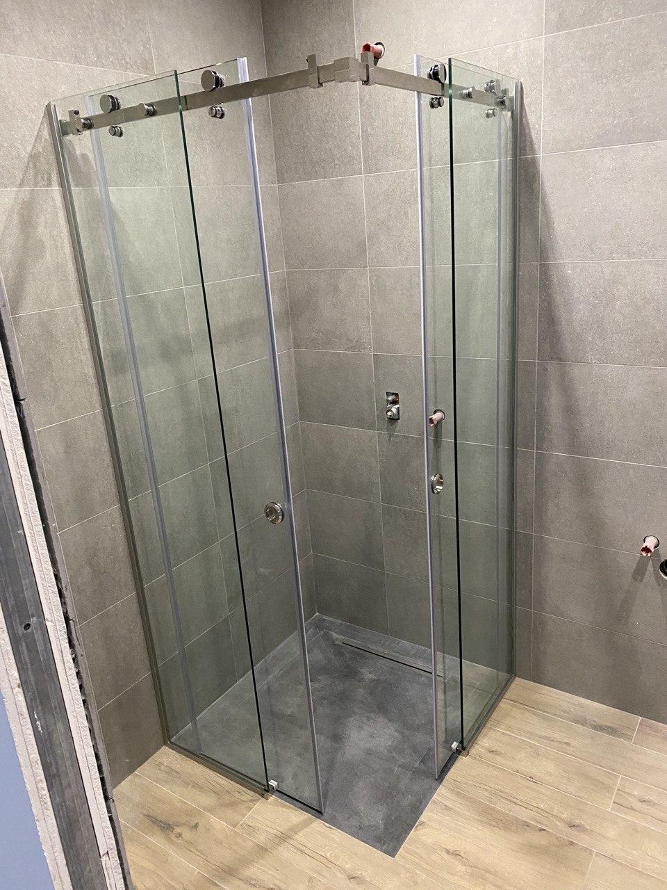 Kabiną prysznicowa narożna z przesuwnymi drzwiami, szklana, z metalowymi okuciami, na tle szarych płytek ściennych i podłogi imitującej drewno.
