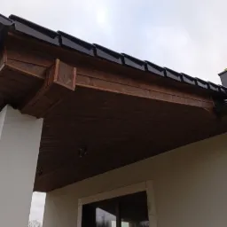 Fragment elewacji domu z widocznym oknem, białym filarem i drewnianym podbitką dachu z czarną dachówką w pochmurny dzień.