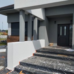 fach-bud - Elewacja domu w trakcie malowania: białe elementy dekoracyjne, szara ściana, zabezpieczone schody folią. Widoczne kolumny i okno z żaluzjami.