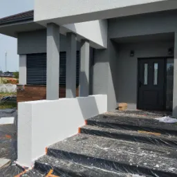 Elewacja domu w trakcie malowania: białe elementy dekoracyjne, szara ściana, zabezpieczone schody folią. Widoczne kolumny i okno z żaluzjami.