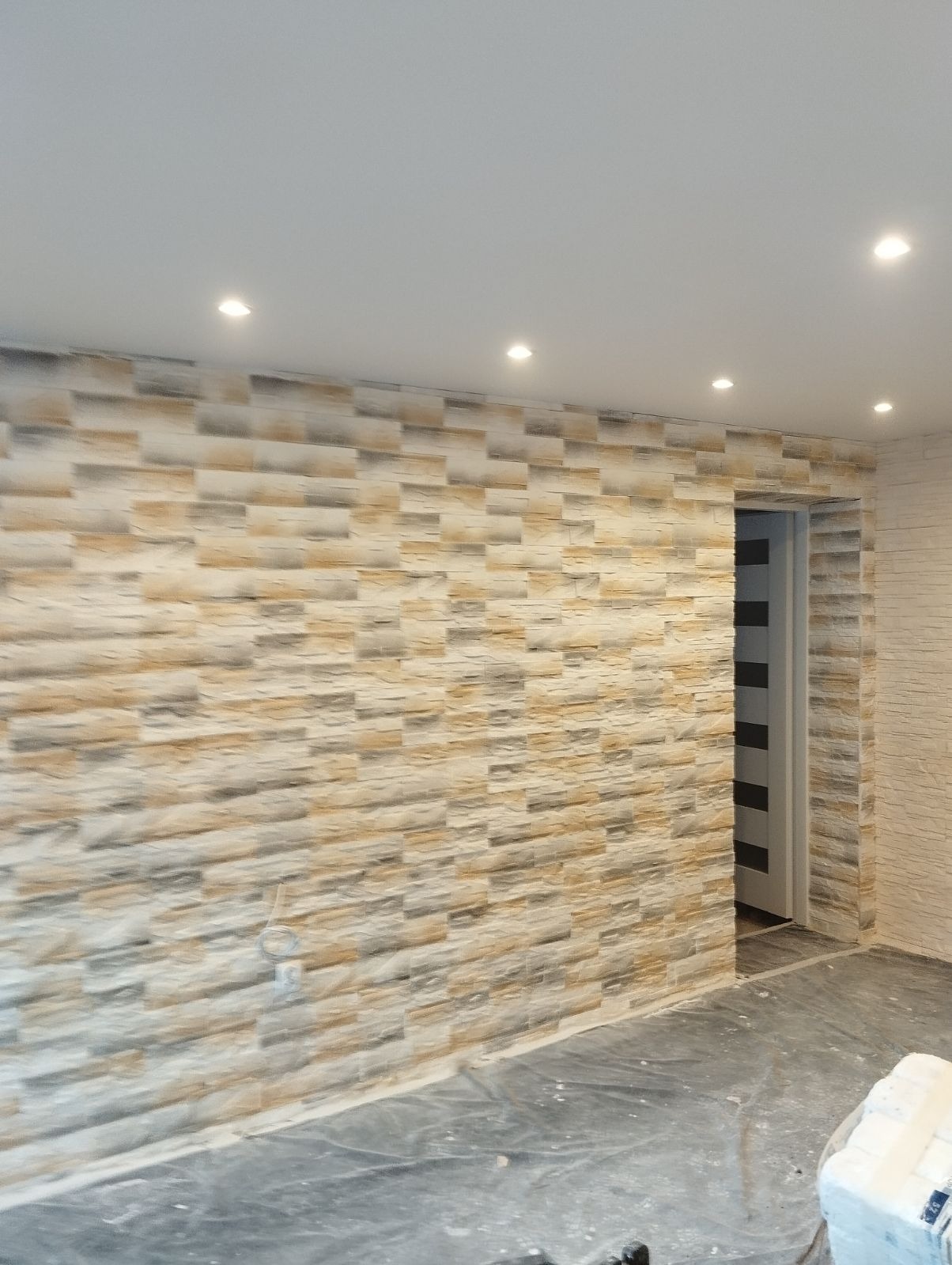 Wnętrze z dekoracyjną ścianą wyłożoną panelami imitującymi kamień w odcieniach beżu i szarości, oświetloną punktowymi lampami sufitowymi, z fragmentem drzwi w tle.
