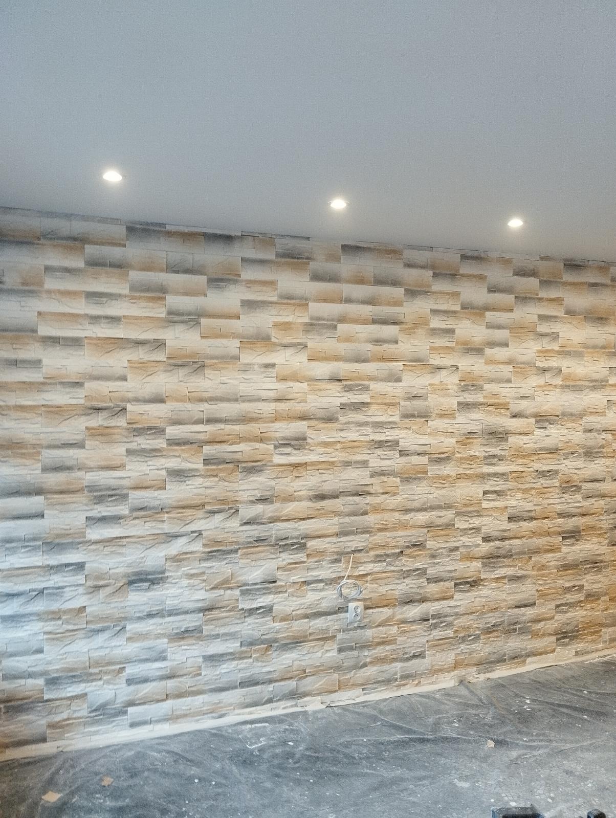 Ściana wykończona panelami imitującymi kamień w odcieniach beżu, szarości i bieli, oświetlona trzema punktowymi lampami sufitowymi, z widocznym wystającym kablem i gniazdkiem elektrycznym,...