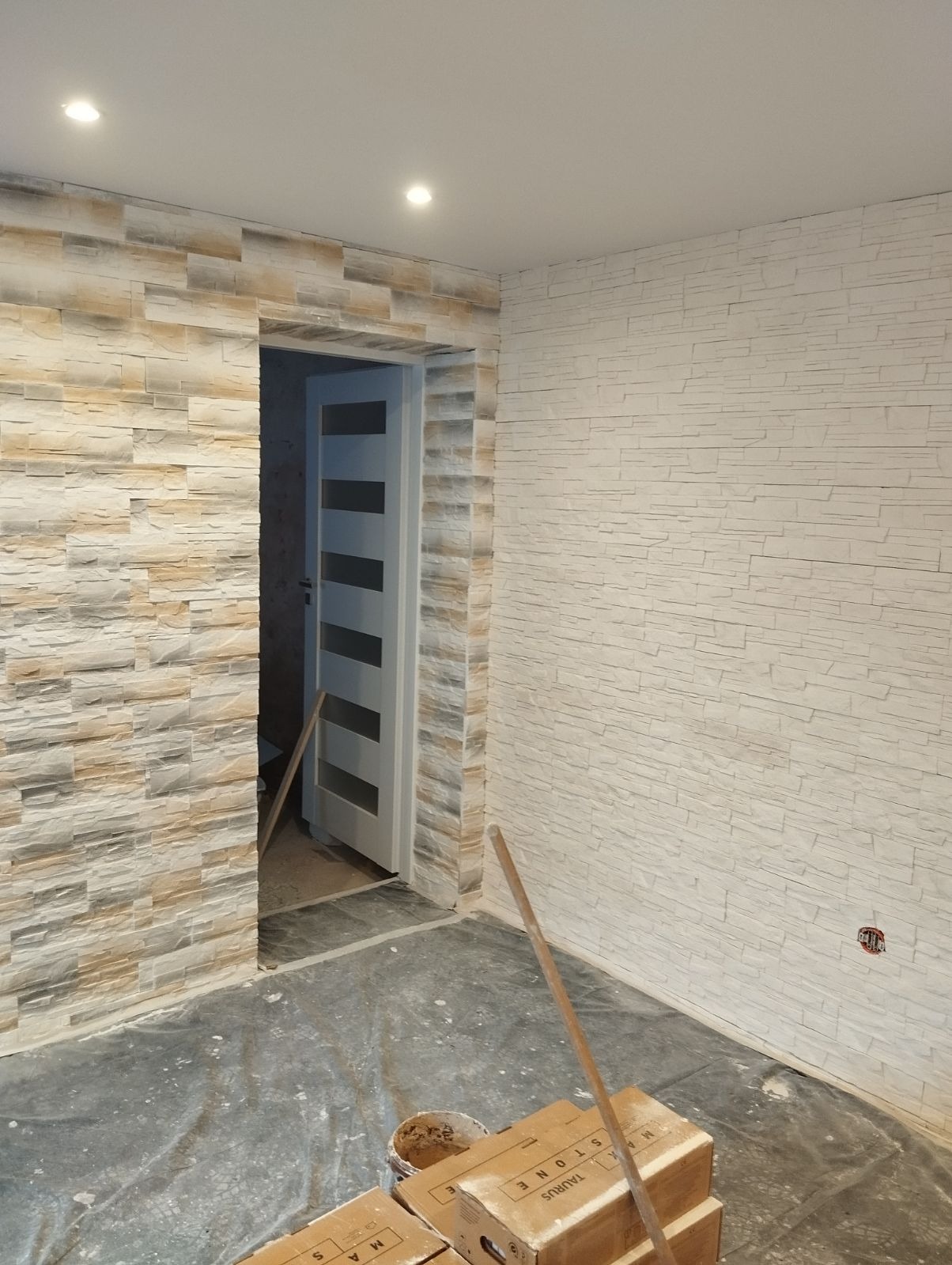Wnętrze pokoju po montażu dekoracyjnej okładziny kamiennej na ścianach, widoczne kartony z okładziną na podłodze zabezpieczonej folią malarską, oraz fragment białych drzwi z przeszkleniami.