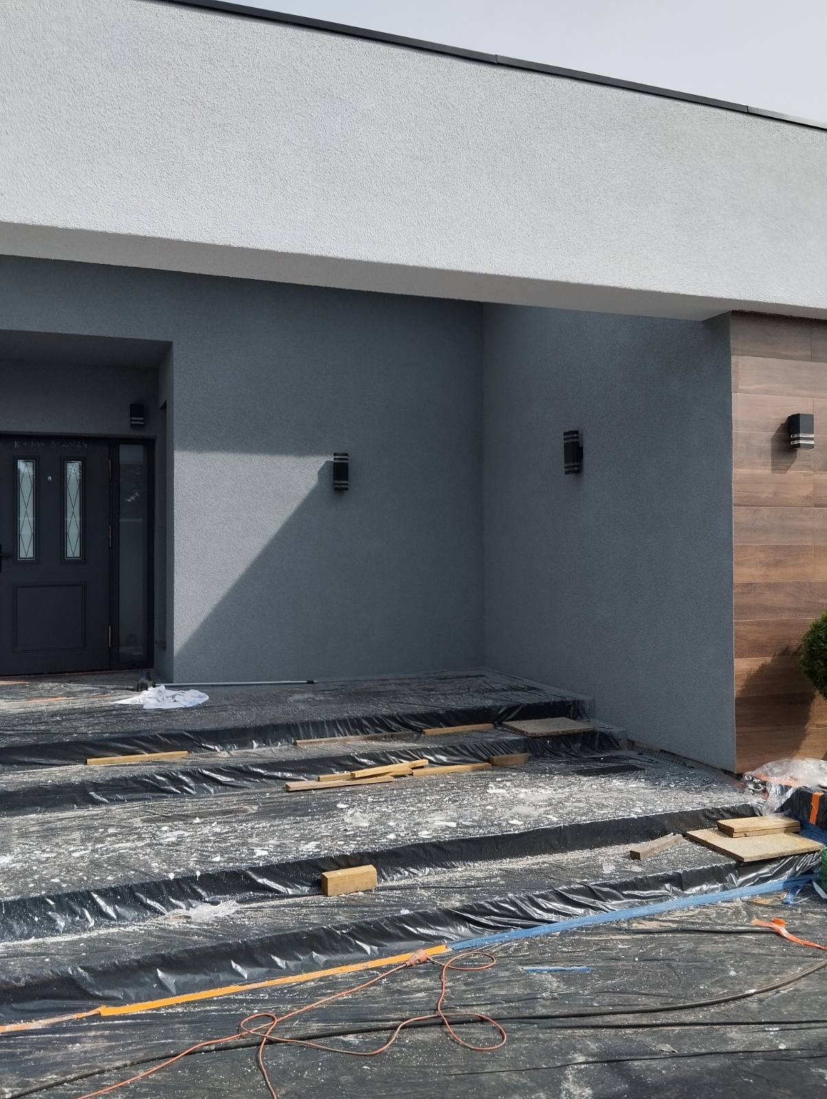 Elewacja domu z szarym tynkiem i drewnianym panelem. Schody zabezpieczone folią, przygotowane do dalszych prac wykończeniowych. Nowoczesne oświetlenie zewnętrzne.