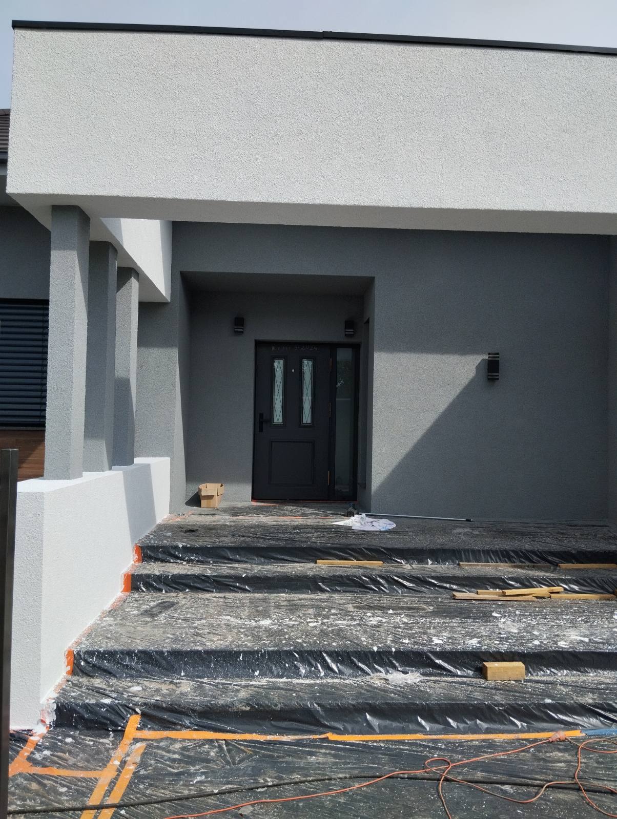 Elewacja domu w trakcie prac: szara ściana, białe elementy, zabezpieczone schody folią malarską. Widoczne lampy zewnętrzne i zarys dachu.