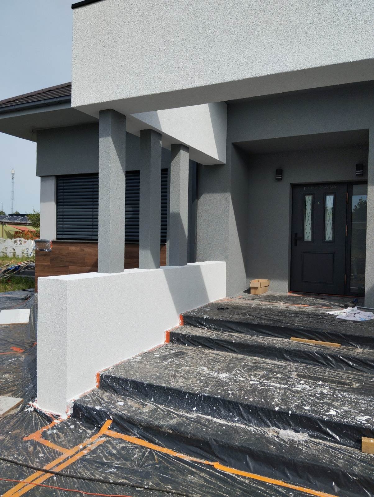 Elewacja domu w trakcie malowania: białe elementy dekoracyjne, szara ściana, zabezpieczone schody folią. Widoczne kolumny i okno z żaluzjami.