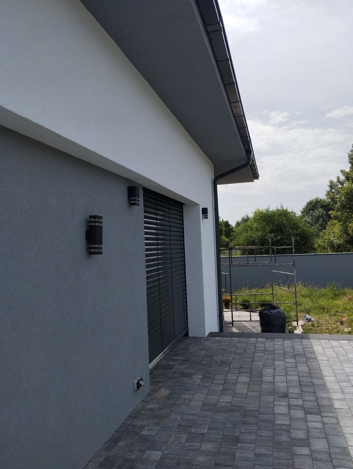 Nowoczesna elewacja domu w odcieniach szarości i bieli, z oknem z żaluzjami zewnętrznymi i oświetleniem elewacyjnym. Widoczne rusztowanie i kostka brukowa.