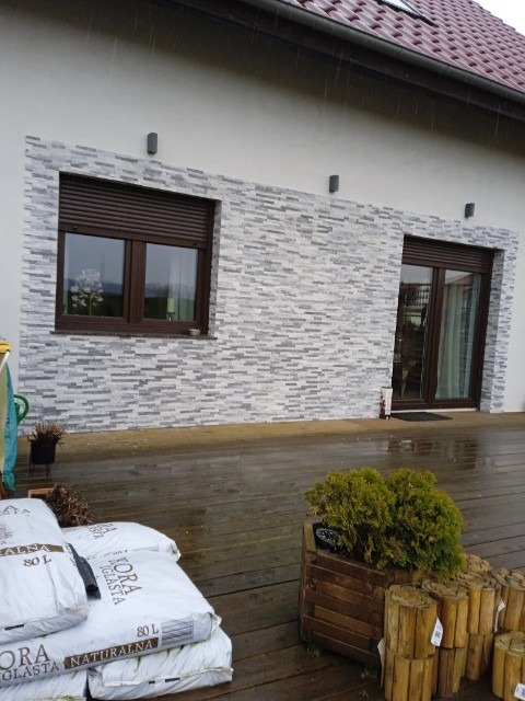 Dom z elewacją z szarej mozaiki, brązowe okna z roletami, drewniany taras i worki ziemi. Widok z zewnątrz w pochmurny dzień.