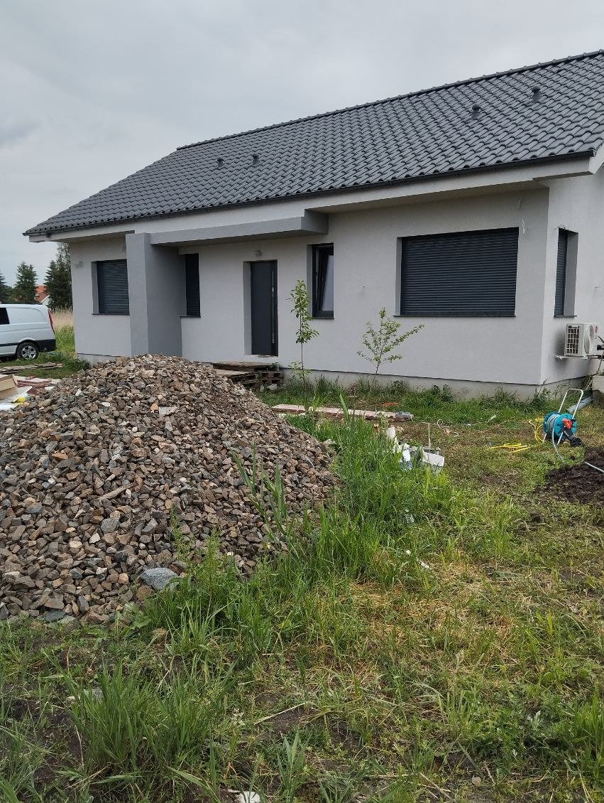 Dom jednorodzinny z szarą elewacją i dachem, z widocznym stosem kamieni na trawniku. Zamontowane rolety zewnętrzne w oknach. W tle biały bus.