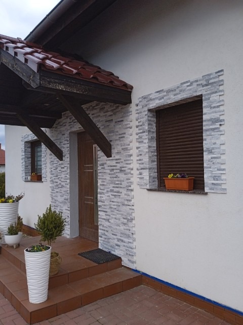 Elewacja domu z szarej cegły dekoracyjnej wokół okna i drzwi. Czerwona dachówka, brązowe rolety okienne i doniczki z kwiatami.