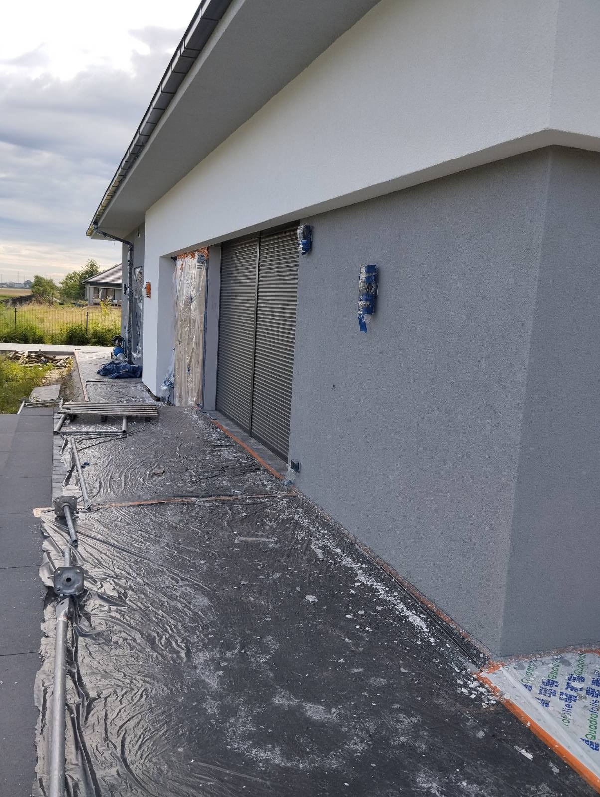 Szara elewacja domu jednorodzinnego w trakcie prac wykończeniowych, zabezpieczona folią, z widocznymi puszkami oświetleniowymi.