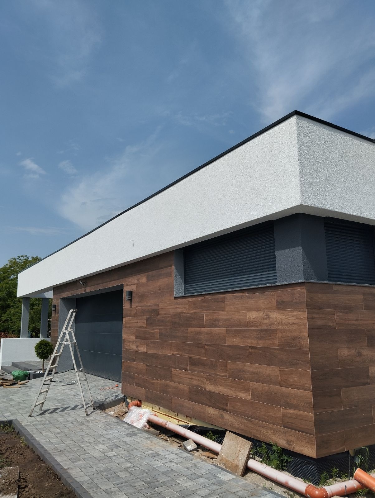 Nowoczesny dom z elewacją imitującą drewno, szarymi roletami i białym, teksturowanym wykończeniem górnej części. Widoczna drabina i fragment chodnika z kostki.