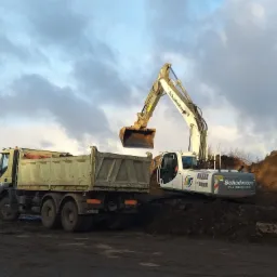 Żółta ciężarówka wywrotka i biała koparka w Koszalinie podczas prac ziemnych, z ciemną ziemią w łyżce koparki gotową do załadunku.