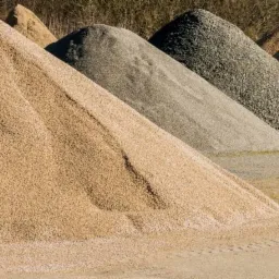 Rzędy stożkowych pryzm różnego kruszywa, od jasnego piasku po ciemny żwir, pod naturalnym oświetleniem.