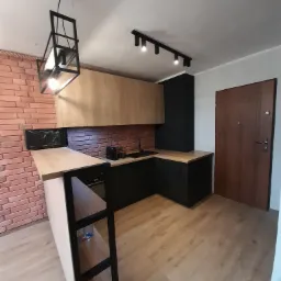 Nowoczesna kuchnia z drewnianymi blatami i czarnymi szafkami, oświetlona wiszącą lampą w stylu industrialnym i szyną z reflektorami, z ceglaną ścianą w tle.