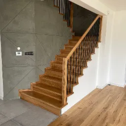 Drewniane schody z czarnymi balustradami, osadzone między dwoma ścianami z dekoracyjnym betonem w geometryczne wzory.