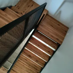 Widok z góry na nowoczesne schody drewniane z metalową balustradą o wzorze siatki, oświetlone naturalnym światłem wpadającym przez szczeliny między stopniami.