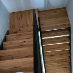 Drewniane schody z czarną, metalową balustradą, widok z góry, ukazujący unikalny rysunek słojów drewna na stopniach.