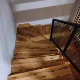 Spiralne schody drewniane z metalową balustradą z siatki, widok z g&oacute;ry, naturalne wykończenie drewna z ciemnymi sękami, oświetlenie z g&oacute;ry tworzące cienie na ścianie.