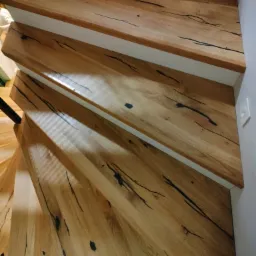 Drewniane schody spiralne z jasnego drewna z ciemnymi żyłkami, białe podstopnice, widok z góry.