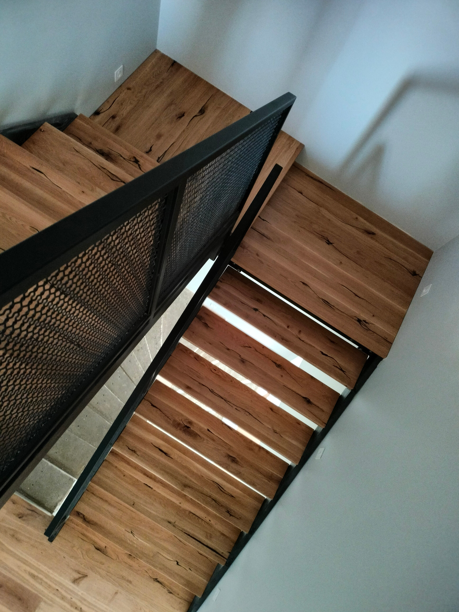 Widok z góry na nowoczesne schody drewniane z metalową balustradą o wzorze siatki, oświetlone naturalnym światłem wpadającym przez szczeliny między stopniami.