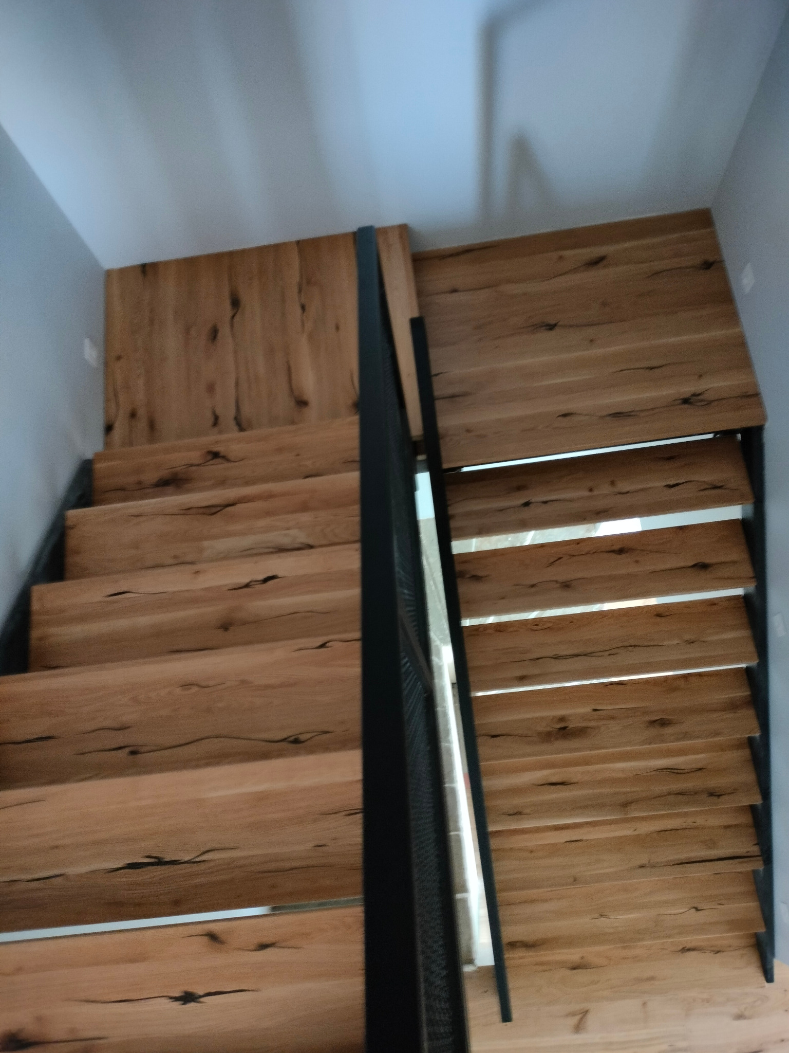 Drewniane schody z czarną, metalową balustradą, widok z góry, ukazujący unikalny rysunek słojów drewna na stopniach.