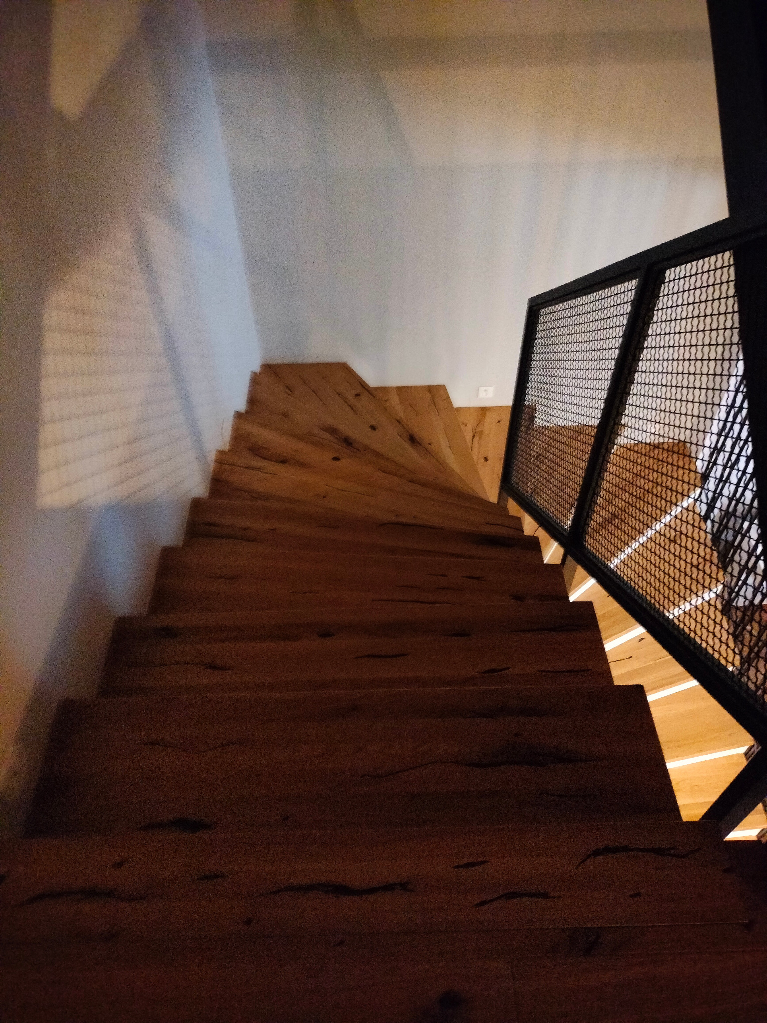 Schody drewniane z metalową balustradą siatkową, widok z góry, z wyraźnym usłojeniem drewna i nieregularnym kształtem stopni.