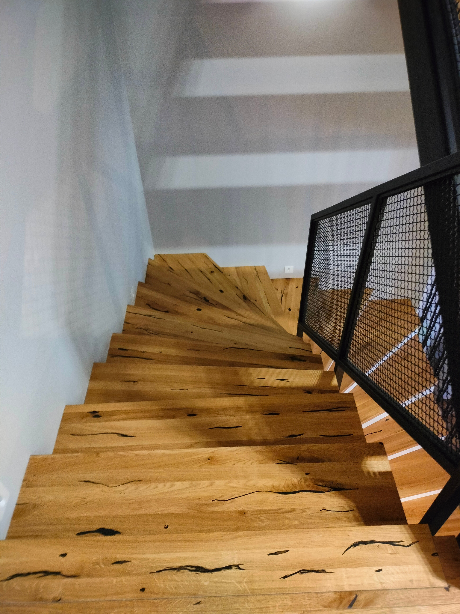 Drewniane schody z wyraźnym usłojeniem i czarnymi żywicznymi wypełnieniami, zabezpieczone metalową balustradą z siatki, widok z góry w kierunku stopni.