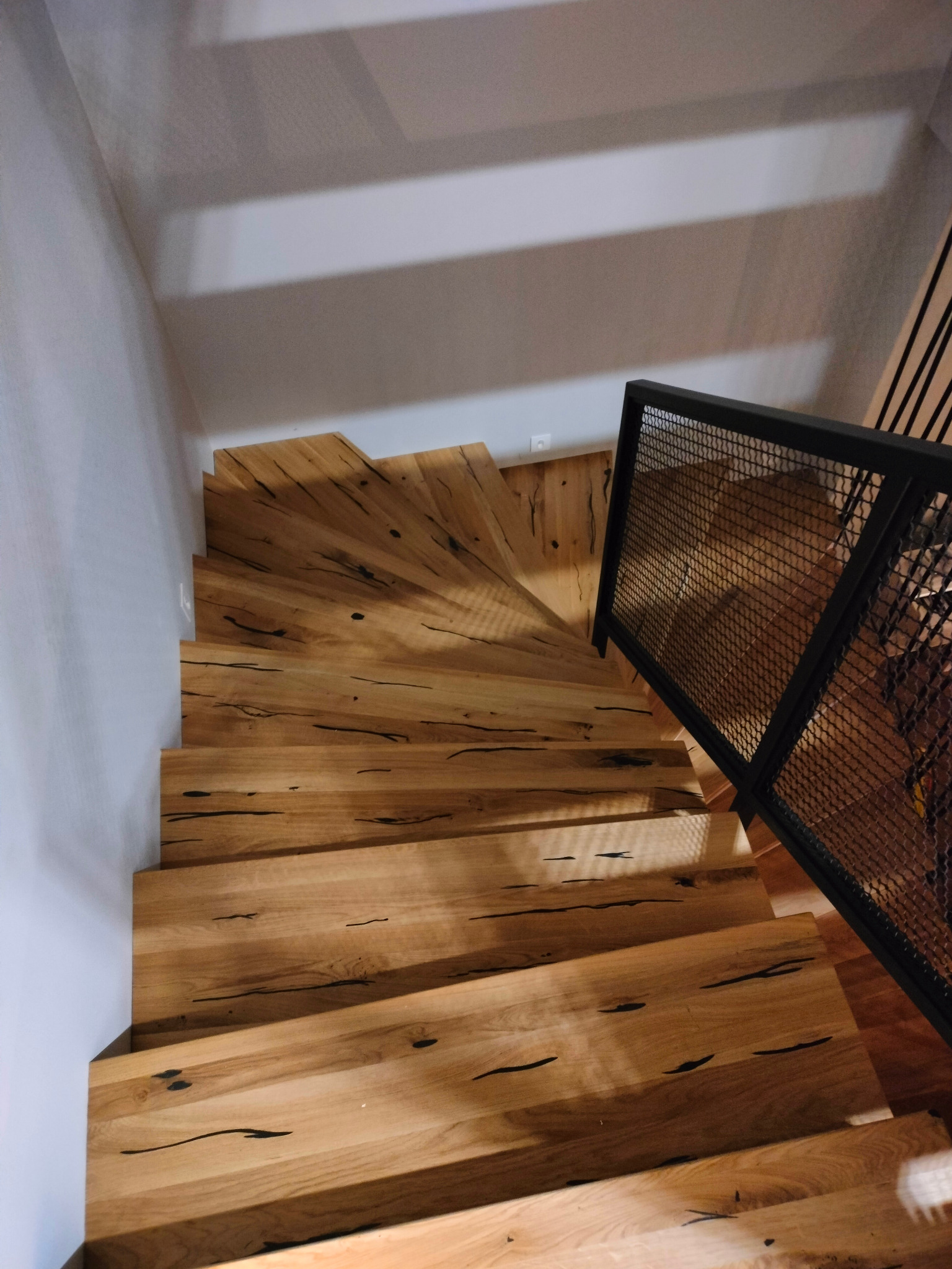 Spiralne schody drewniane z metalową balustradą z siatki, widok z góry, naturalne wykończenie drewna z ciemnymi sękami, oświetlenie z góry tworzące cienie na ścianie.