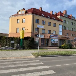 Widok zewnętrzny trzypiętrowego budynku z czerwonym dachem, w którym znajdują się biura i lokale usługowe, w tym prawdopodobnie kancelaria prawna, co sugeruje tabliczka z logiem na fasadzie.