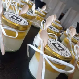 Rząd słoiczków z deserem, warstwa białej pianki i żółtego musu, złote wieczka z logo, drewniane łyżeczki i białe wstążki, przygotowane na catering.