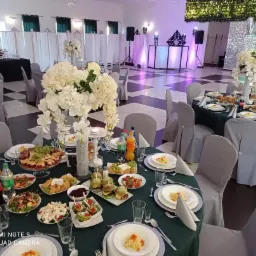 Uroczystość osiemnastych urodzin: sala bankietowa z okrągłymi stołami nakrytymi zielonymi obrusami, dekoracje z białych kwiatów, zastawa stołowa, napoje, przekąski, parkiet w biało-czarną...