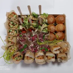 Drewniana skrzynka wypełniona r&oacute;żnorodnymi przekąskami: tortille z kurczakiem i warzywami, mini sałatki z drewnianymi łyżeczkami, desery w kubeczkach z granatem i miętą oraz mini burgery zawinięte...