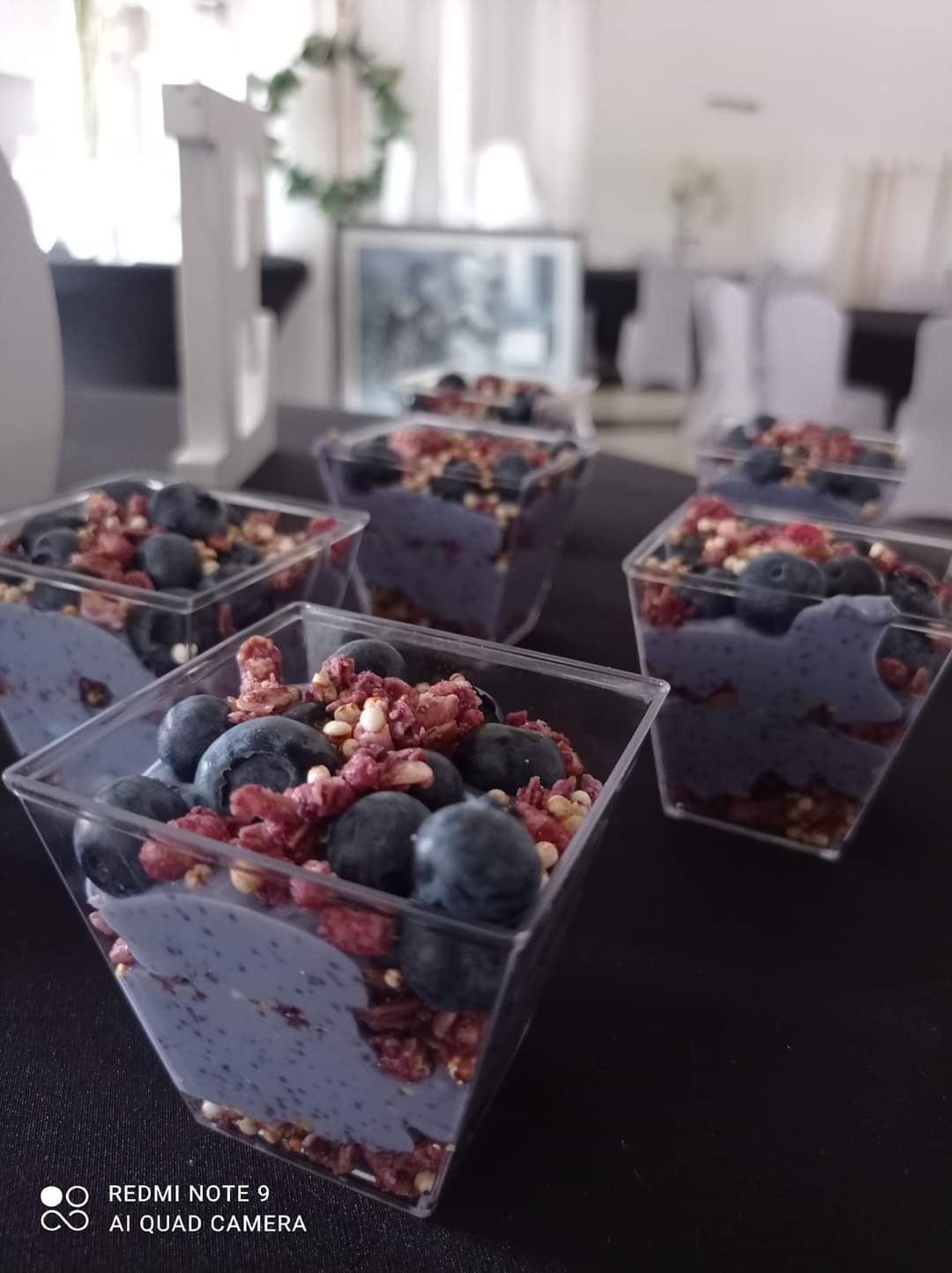 Deser w przezroczystych pojemnikach: granolą, niebieskim kremem i borówkami na czarnym stole, w tle dekoracje sali.