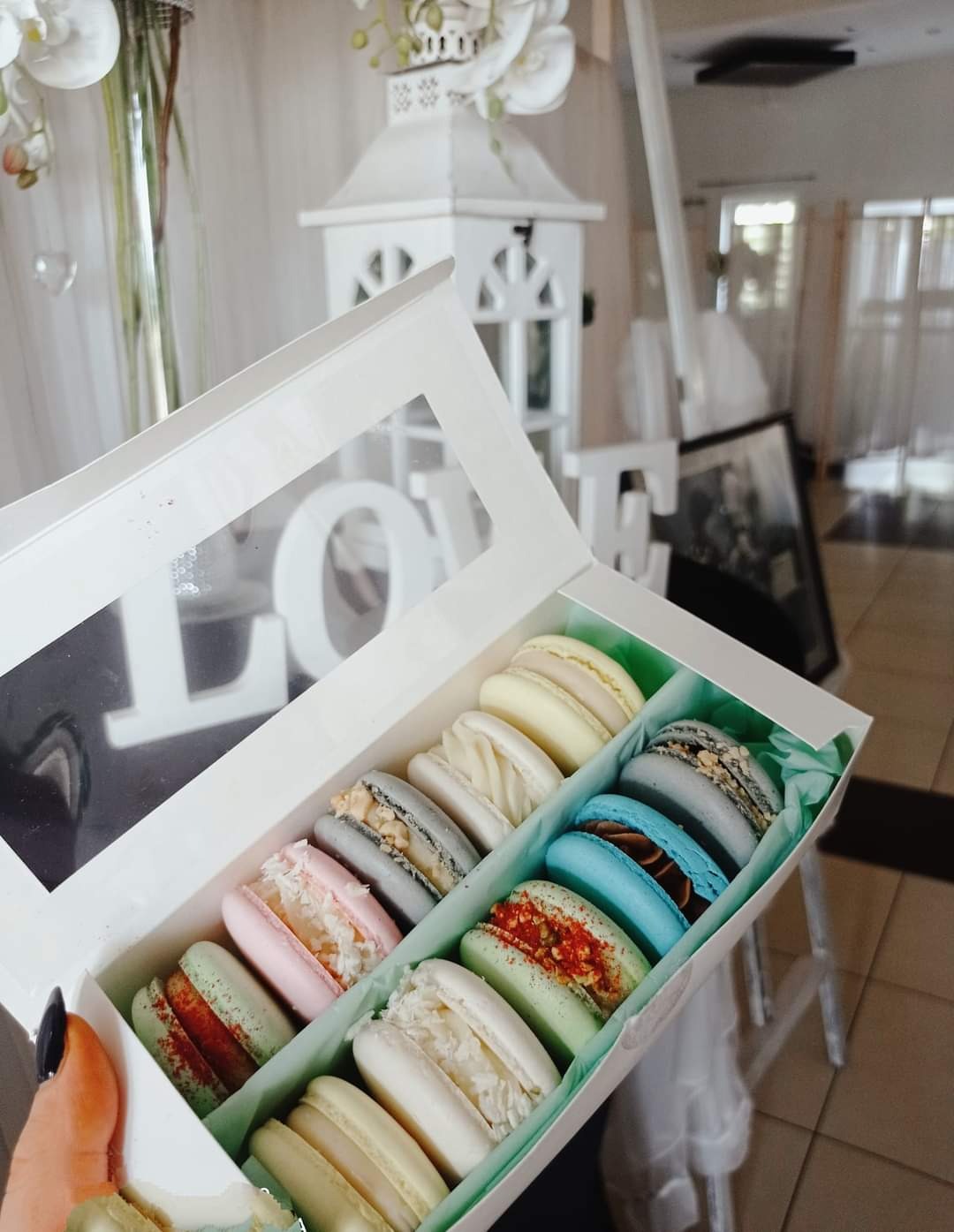 Otwarty kartonik z pastelowymi makaronikami o różnych smakach, ułożonymi na zielonej bibułce; w tle dekoracje ślubne, białe lampiony i kwiaty.