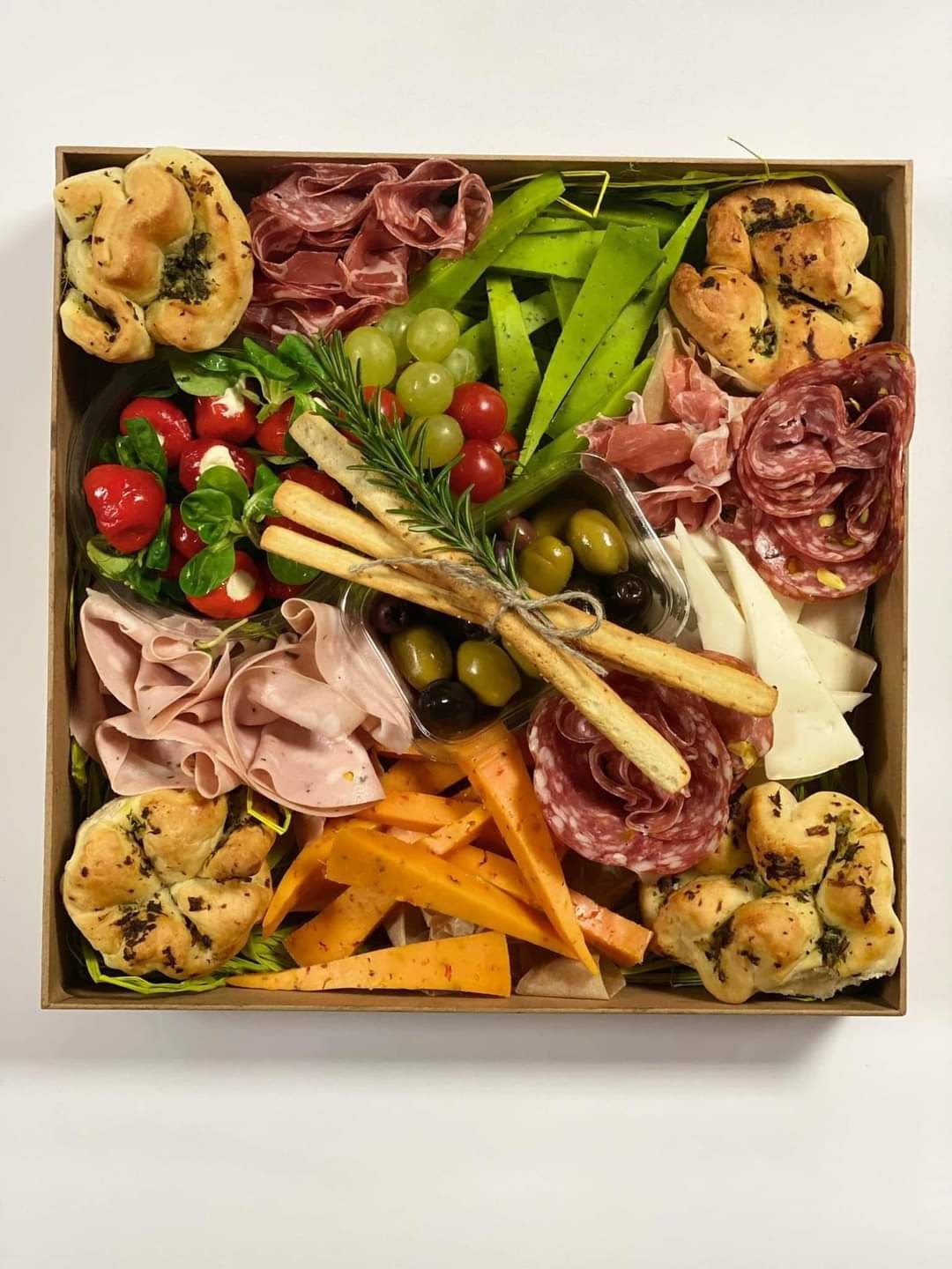 Kartonowe pudełko wypełnione różnorodnymi przekąskami: salami, szynka, oliwki, sery w różnych kolorach, grissini związane sznurkiem, pieczywo ziołowe, papryczki nadziewane serem i winogrona...