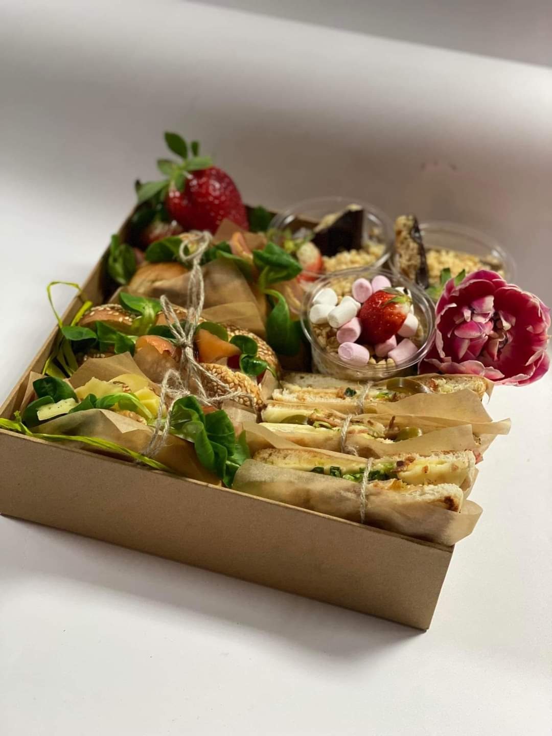 Kartonowe pudełko z różnorodnym cateringiem: kanapki w papierze kraft przewiązane sznurkiem, mini desery w przezroczystych kubeczkach, dekorowane truskawkami i kwiatem tulipana.