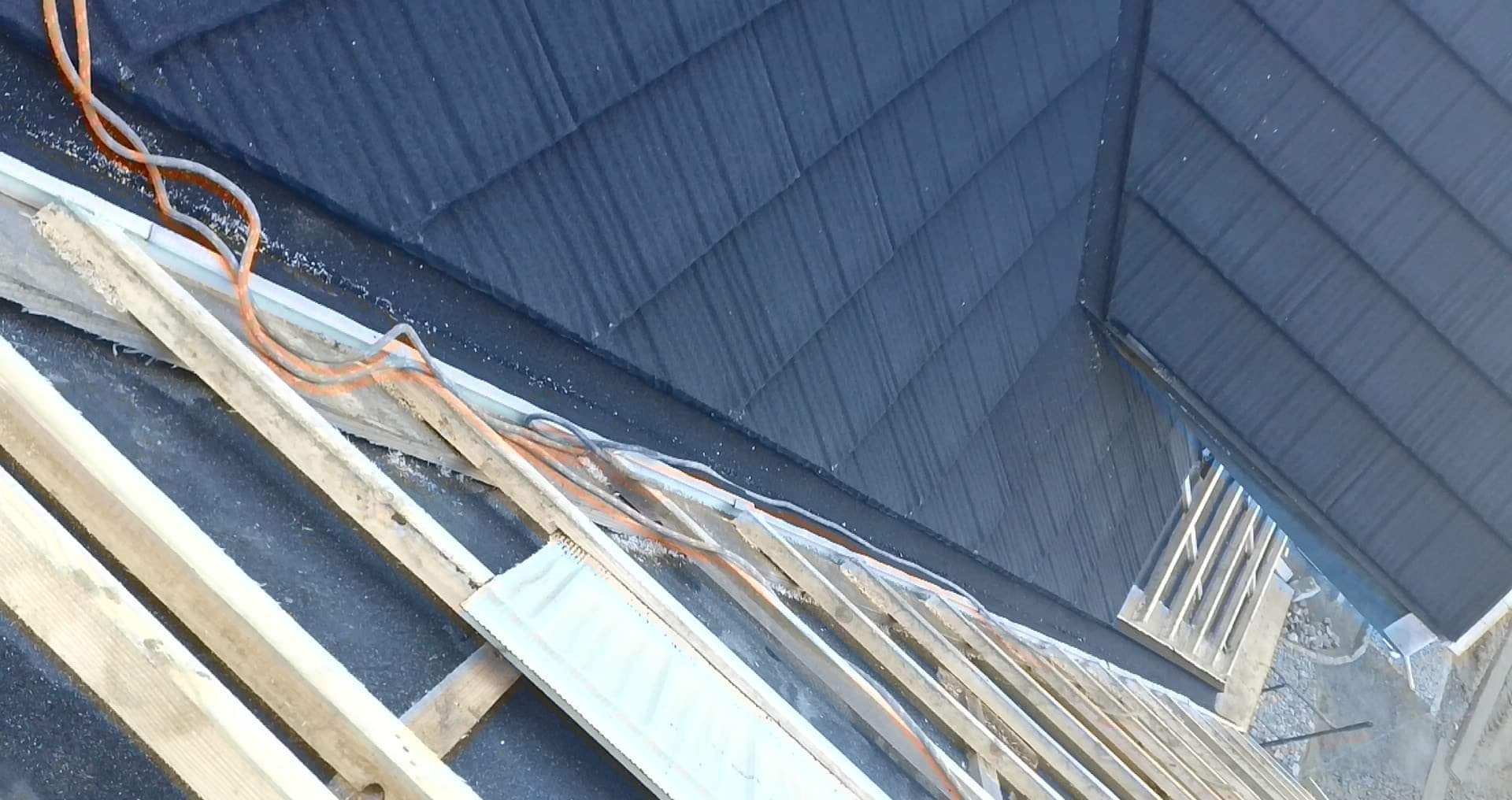 Widok z góry na konstrukcję dachu w trakcie budowy, z widoczną nową, ciemną blachodachówką i elementami drewnianej więźby, oraz wystającymi kablami elektrycznymi.