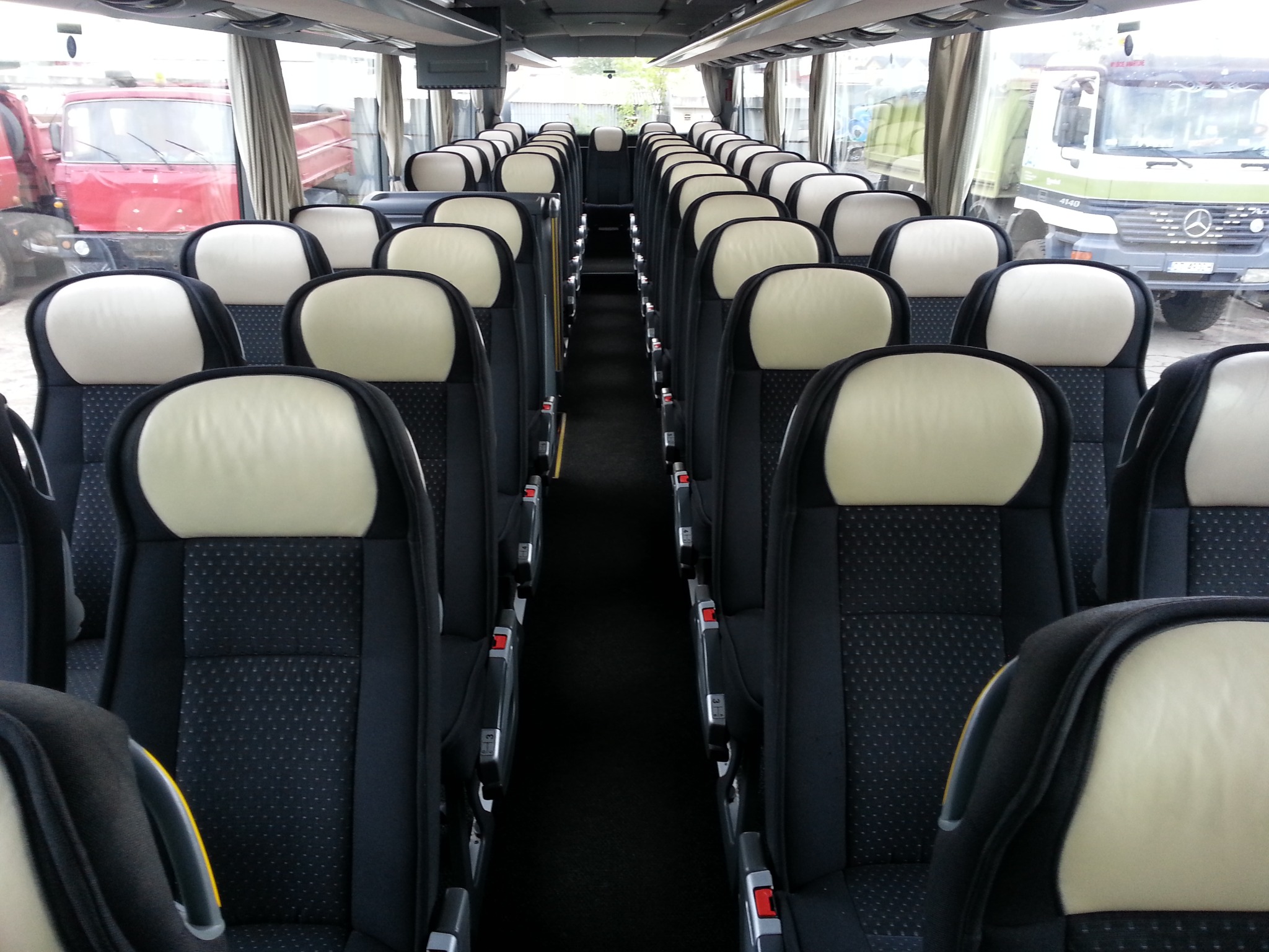 Wnętrze autobusu z rzędami czarnych foteli z beżowymi zagłówkami, widok na okna z zaparkowanymi ciężarówkami na zewnątrz.