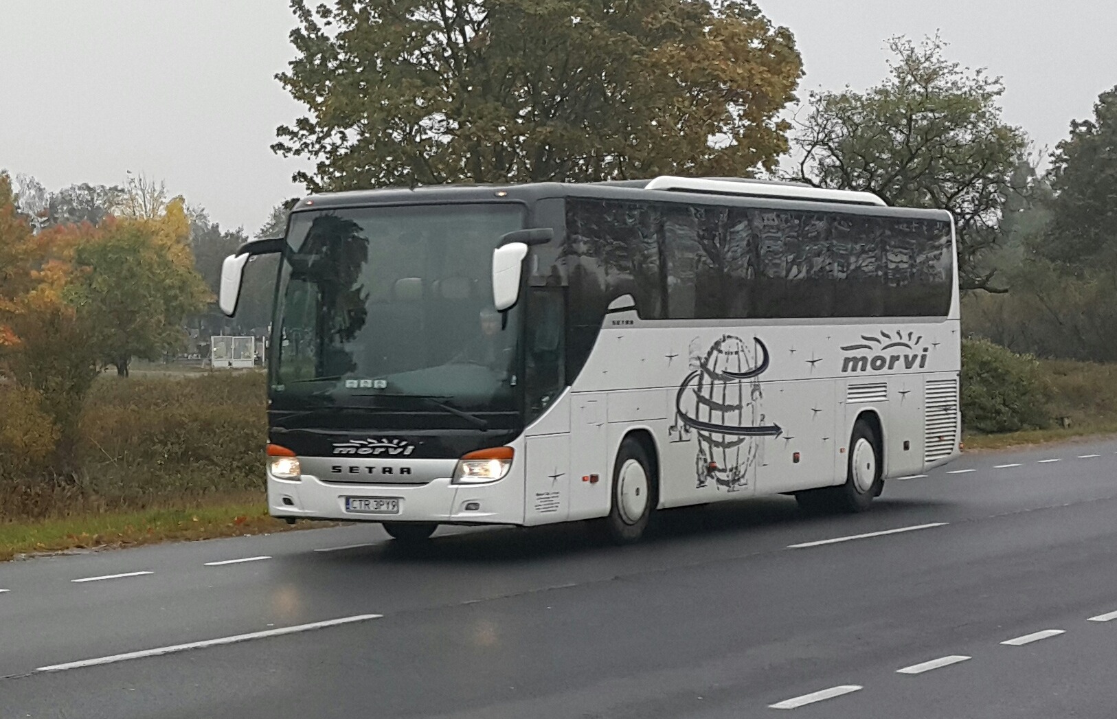Biały autokar marki Setra z grafiką kuli ziemskiej i logo 'morvi' na boku, jadący po mokrej asfaltowej drodze w pochmurny dzień.