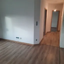 Wykończone wnętrze: świeżo ułożone panele podłogowe z widocznymi listwami przypodłogowymi, gładkie białe ściany z zamontowanymi gniazdkami elektrycznymi i włącznikami, otwarte drzwi do kolejnych...