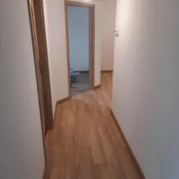 Wnętrze korytarza z drewnopodobną podłogą, widoczne dwie ościeżnice z zamontowanymi drzwiami, surowe wykończenie ścian, żarówka na kablu zwisająca z sufitu.