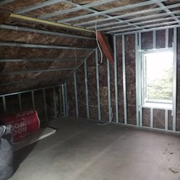 WNB Decor - Poddasze w trakcie prac izolacyjnych: widoczne metalowe profile konstrukcyjne, brązowa wełna mineralna, częściowo otwarte okno z widokiem na zewnątrz i rolka materiału izolacyjnego Rockwool...