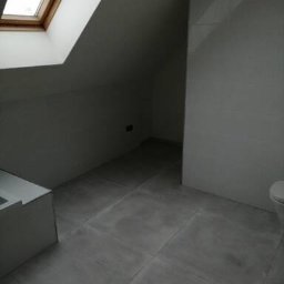 WNB Decor - Szara łazienka na poddaszu z oknem dachowym i dużymi szarymi płytkami na podłodze i ścianach, widoczny fragment wanny i toalety.